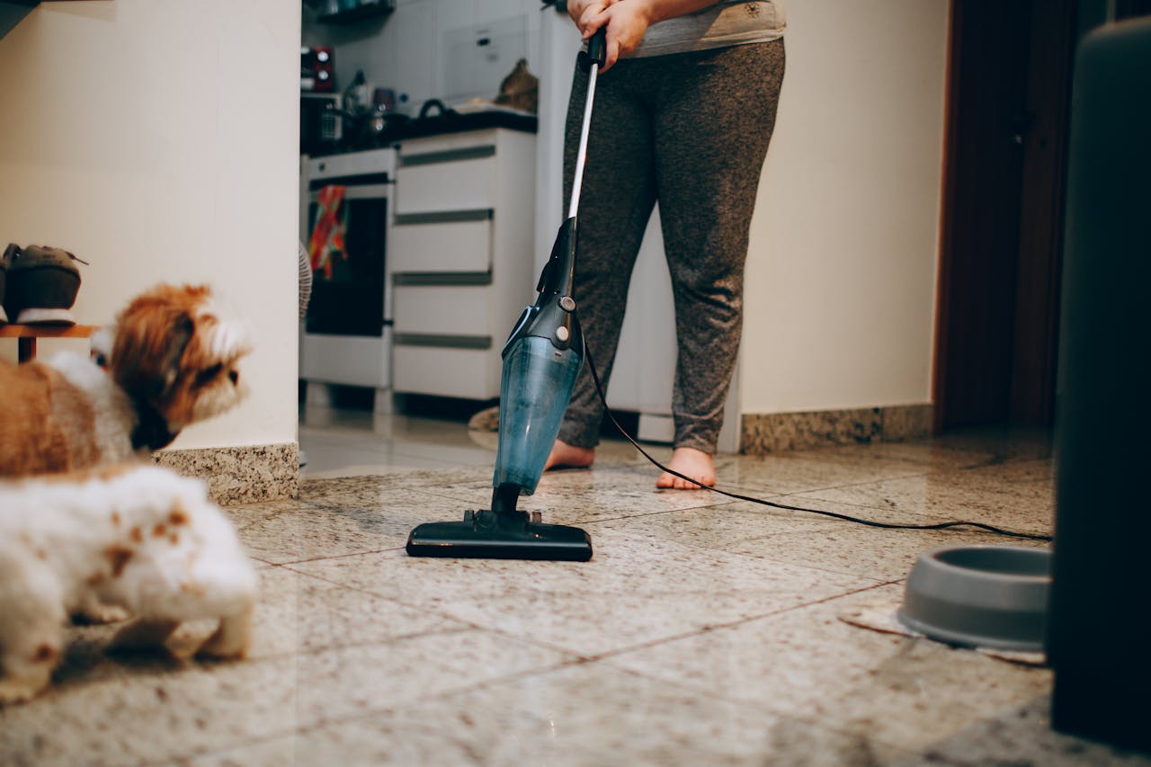 hero-img-02 A woman cleaning the floor with a vacuum cleaner while a dog watches attentively. Indoor home setting.